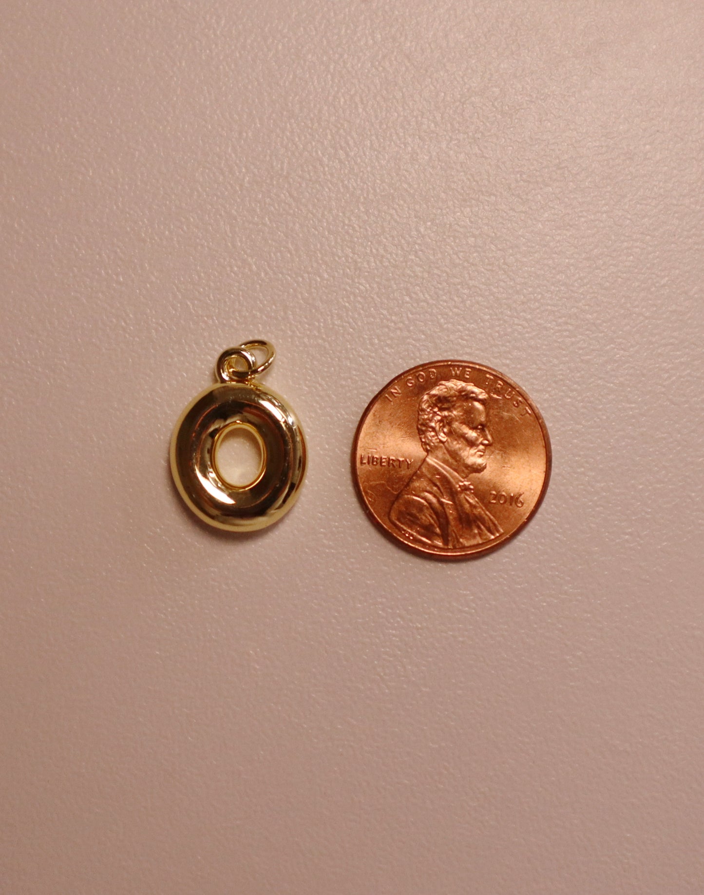 Gold letter pendant next to a penny on a beige background