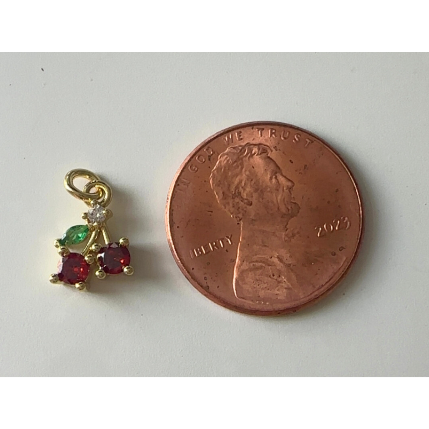 Gold cherry charm with red and green stones next to a penny on a white background