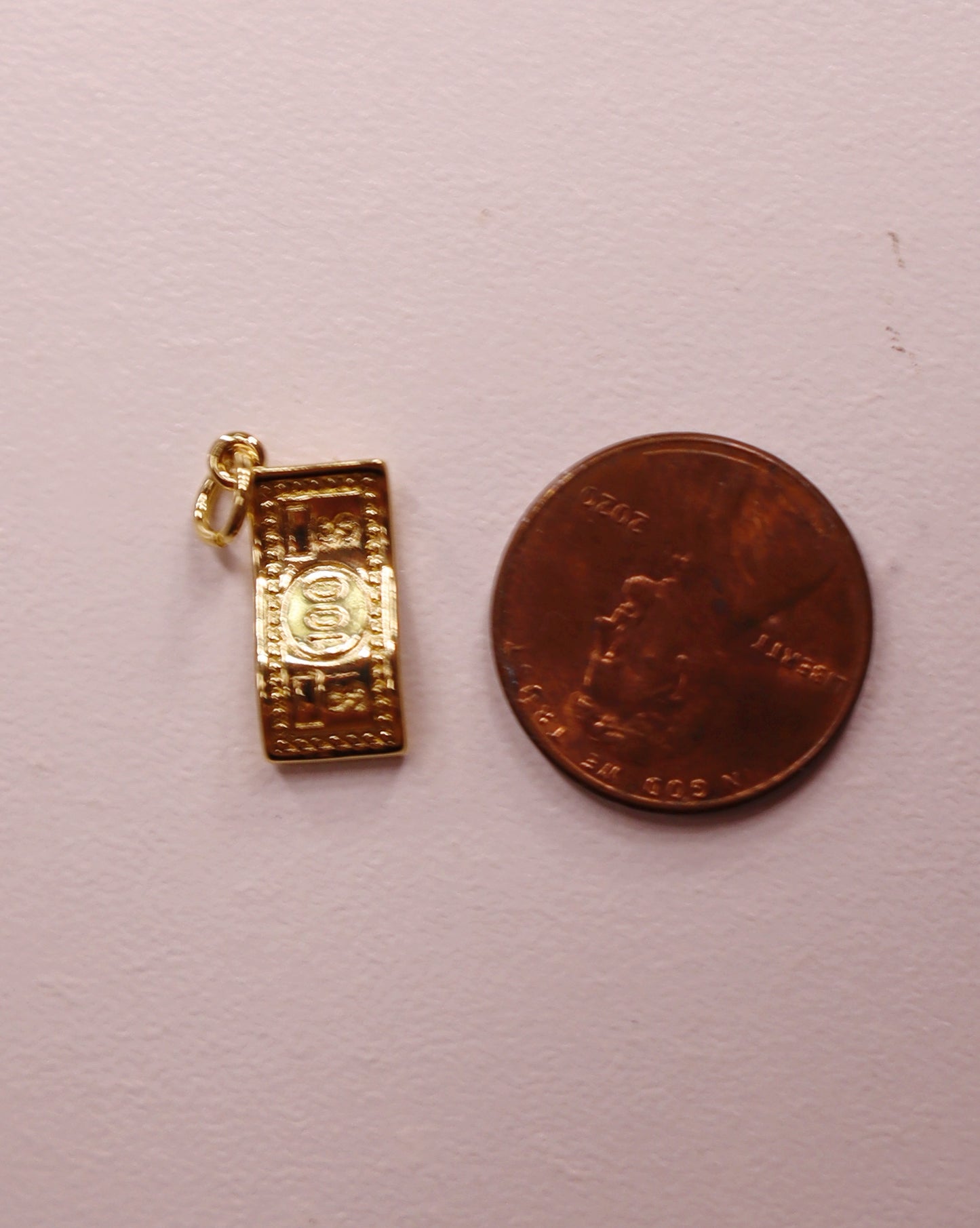 Gold 100 dollar bill pendant next to a penny on a light pink background