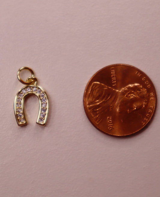 Gold horseshoe charm with clear stones next to a penny on a pink background