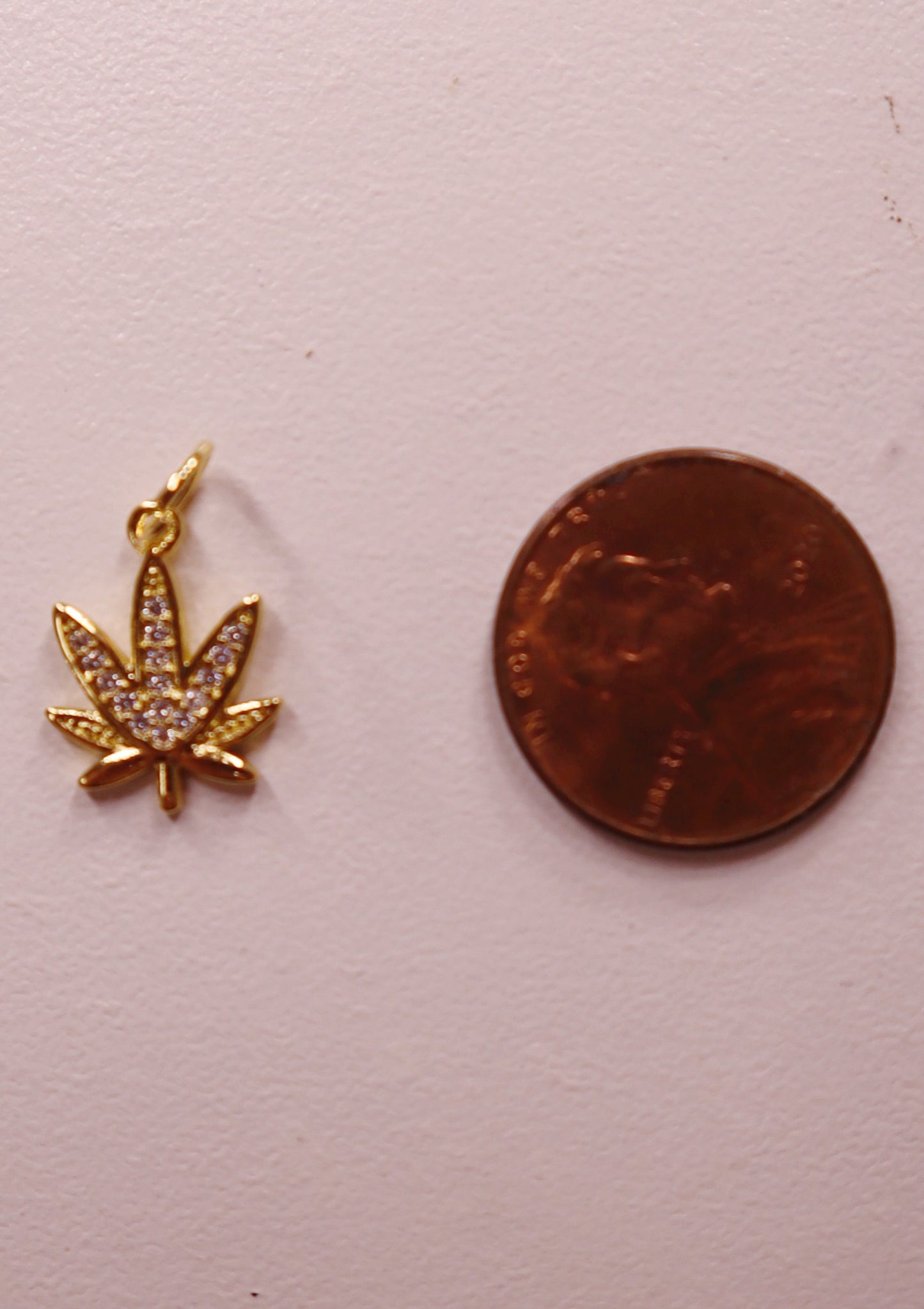 Gold marijuana leaf charm next to a penny on a light pink background