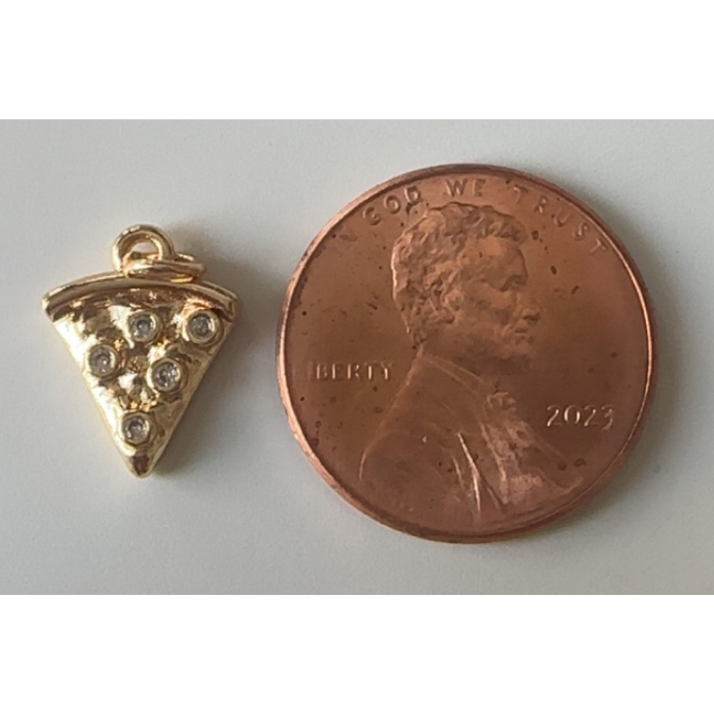 Gold pizza slice charm next to a penny on a light gray background