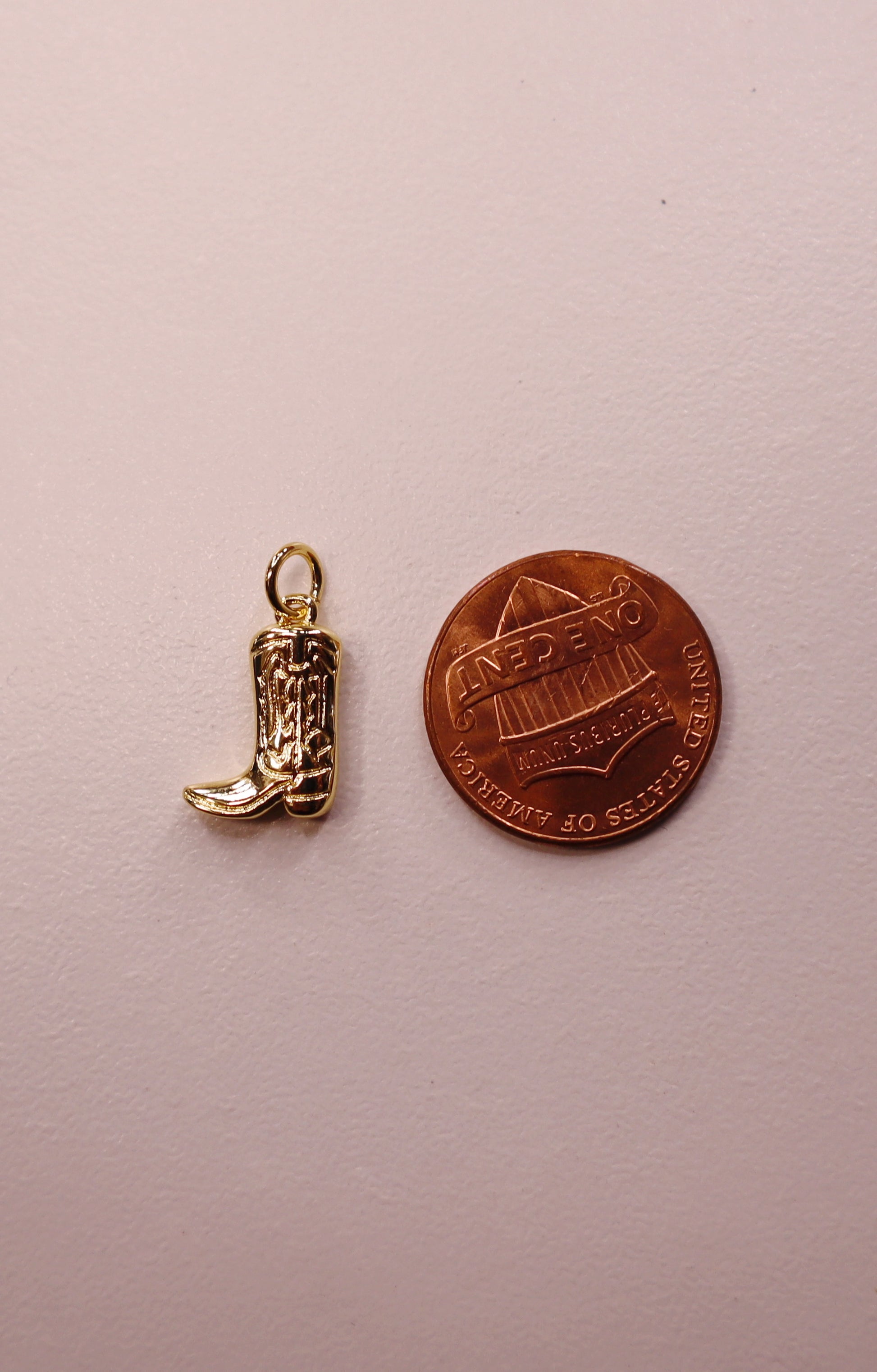 Gold cowboy boot charm next to a copper penny on a pink background