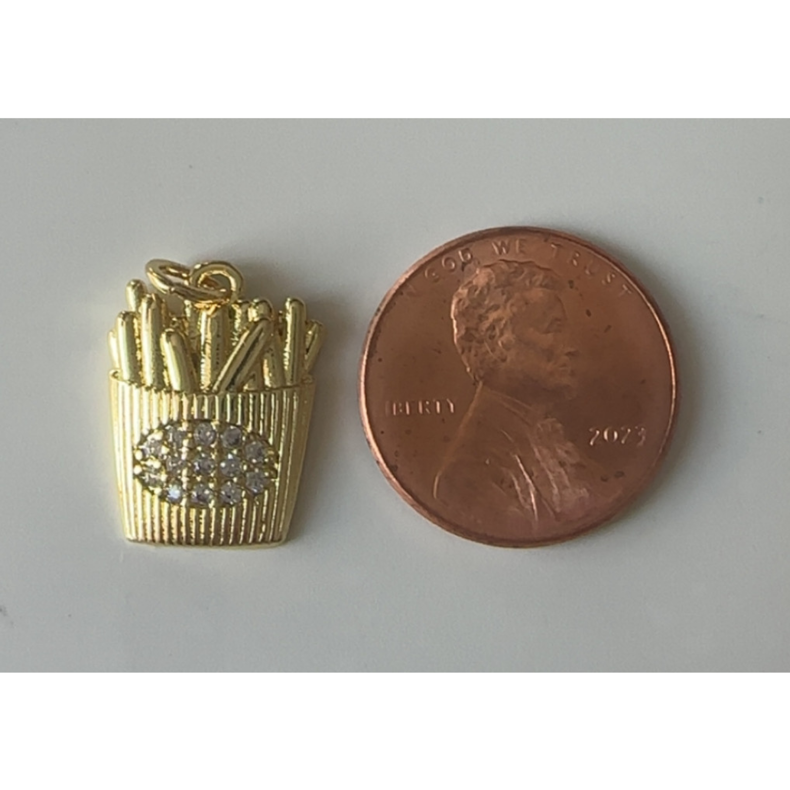 Gold French fry charm next to a penny on a gray background