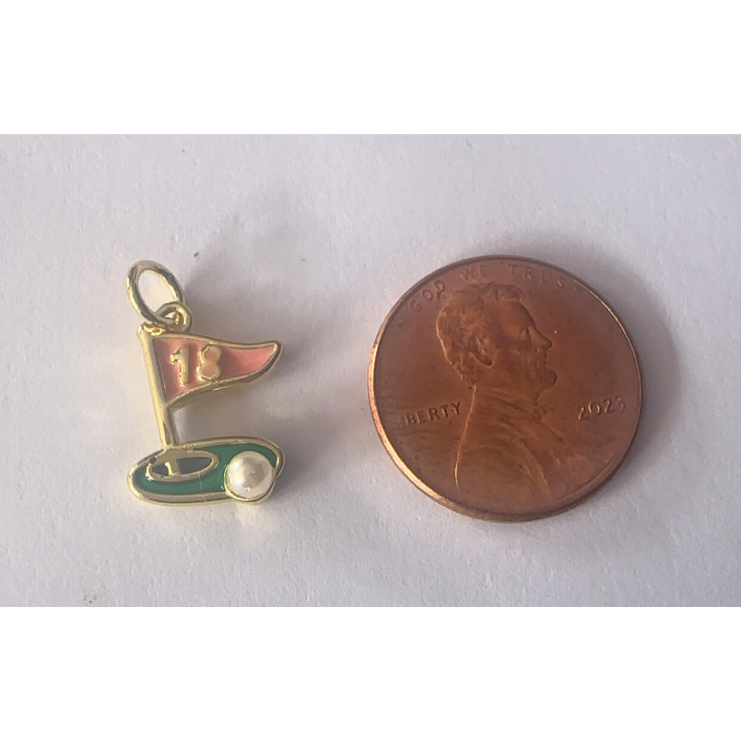 Gold and green golf flag charm next to a penny on a white background