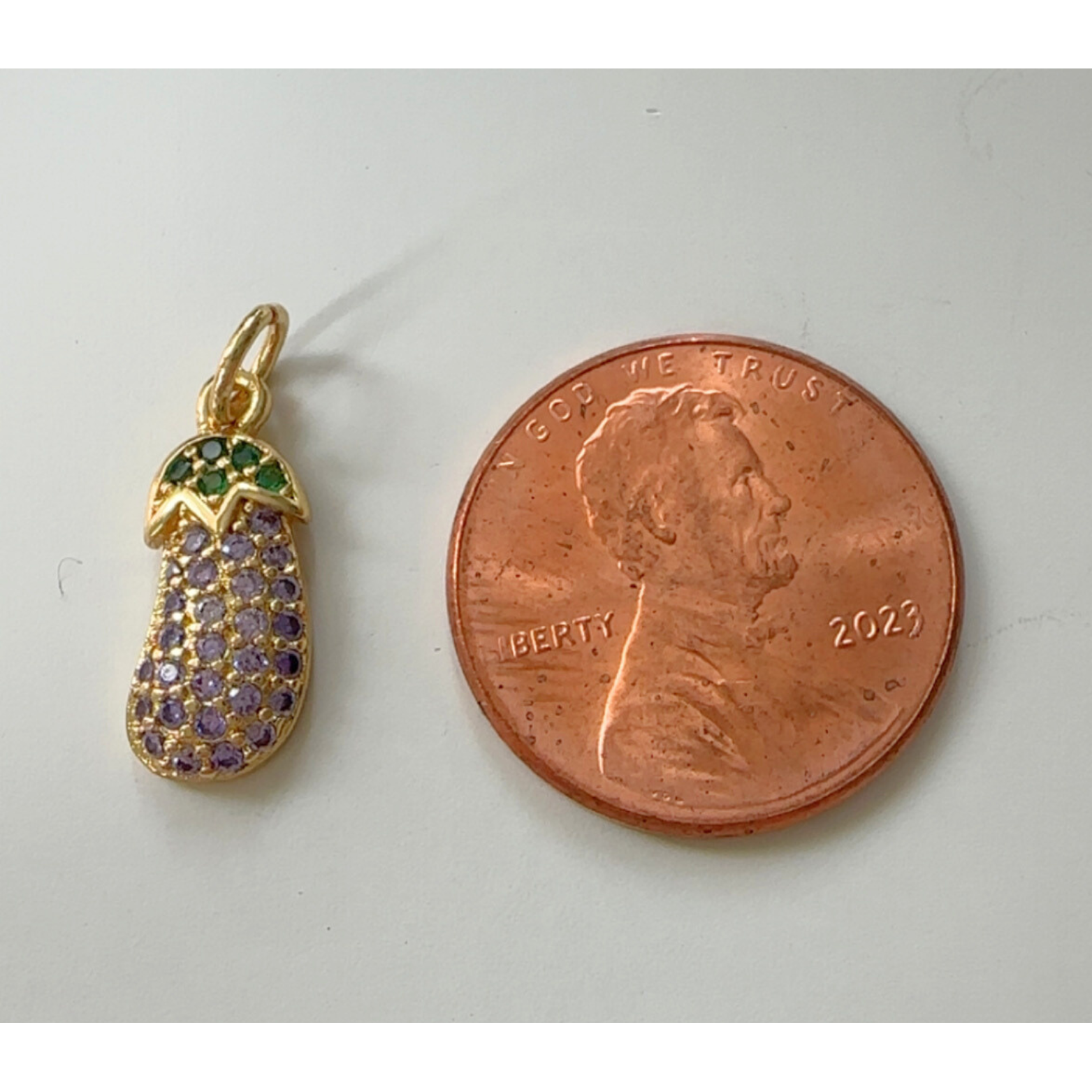 Gold eggplant pendant with gemstones next to a penny on a white background