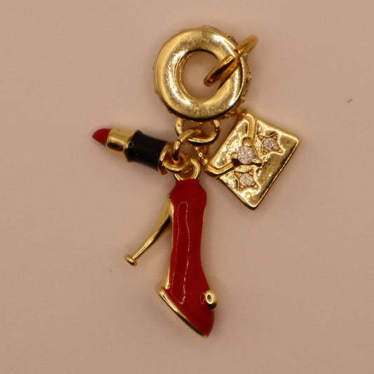Gold charm with a red high heel, lipstick, and heart-shaped diamond pendant on a beige background.