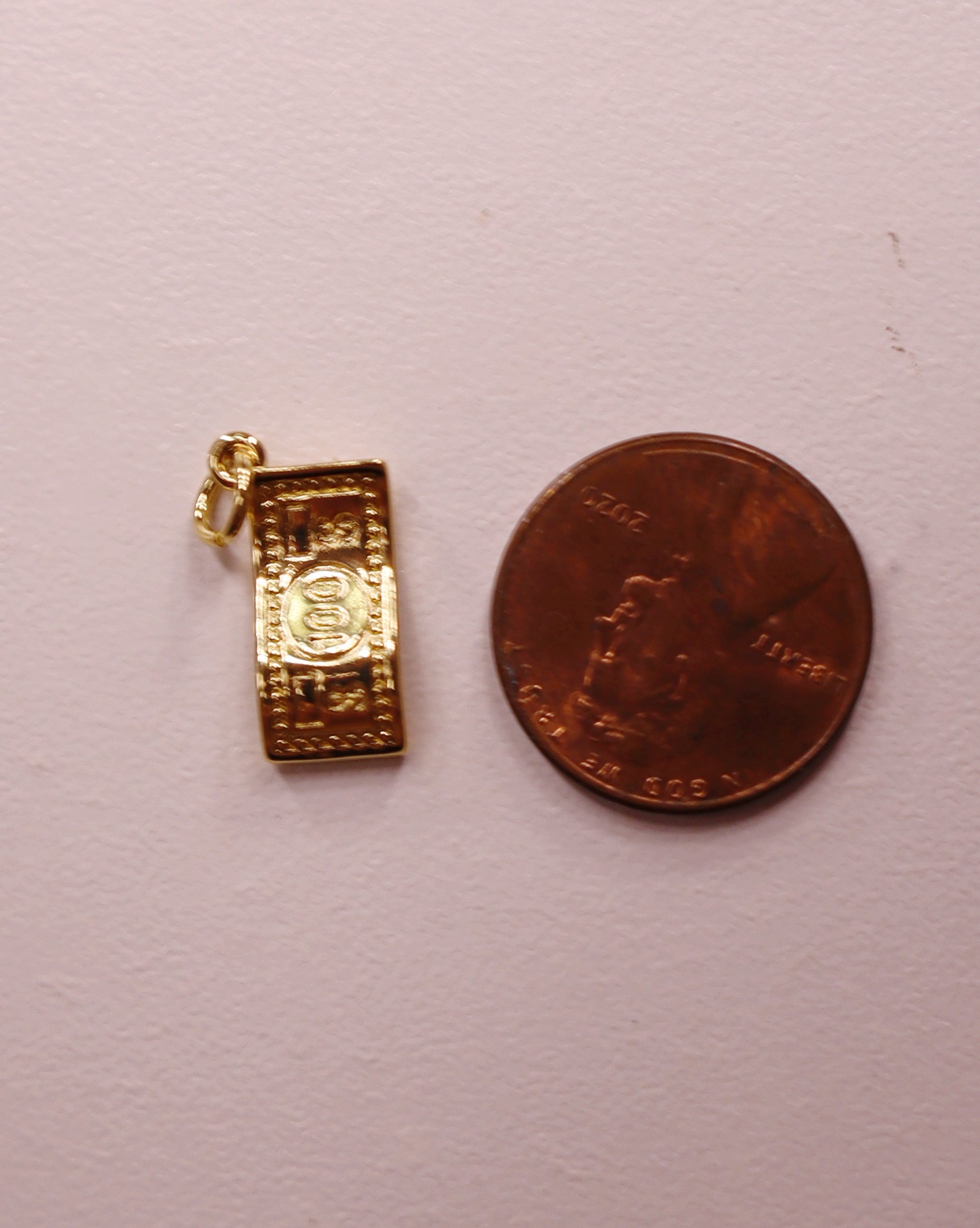 Gold 100 dollar bill pendant next to a penny on a light pink background