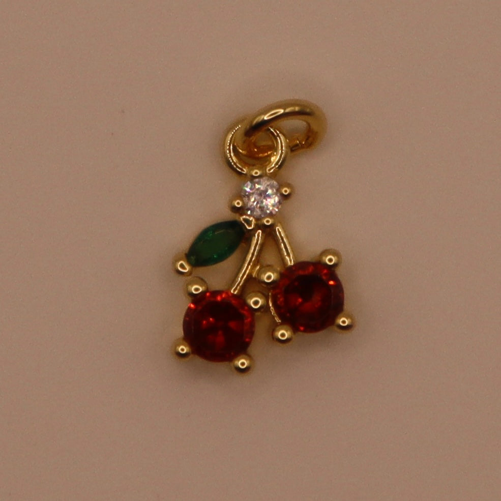 Gold cherry charm with red stones and a green leaf on a beige background