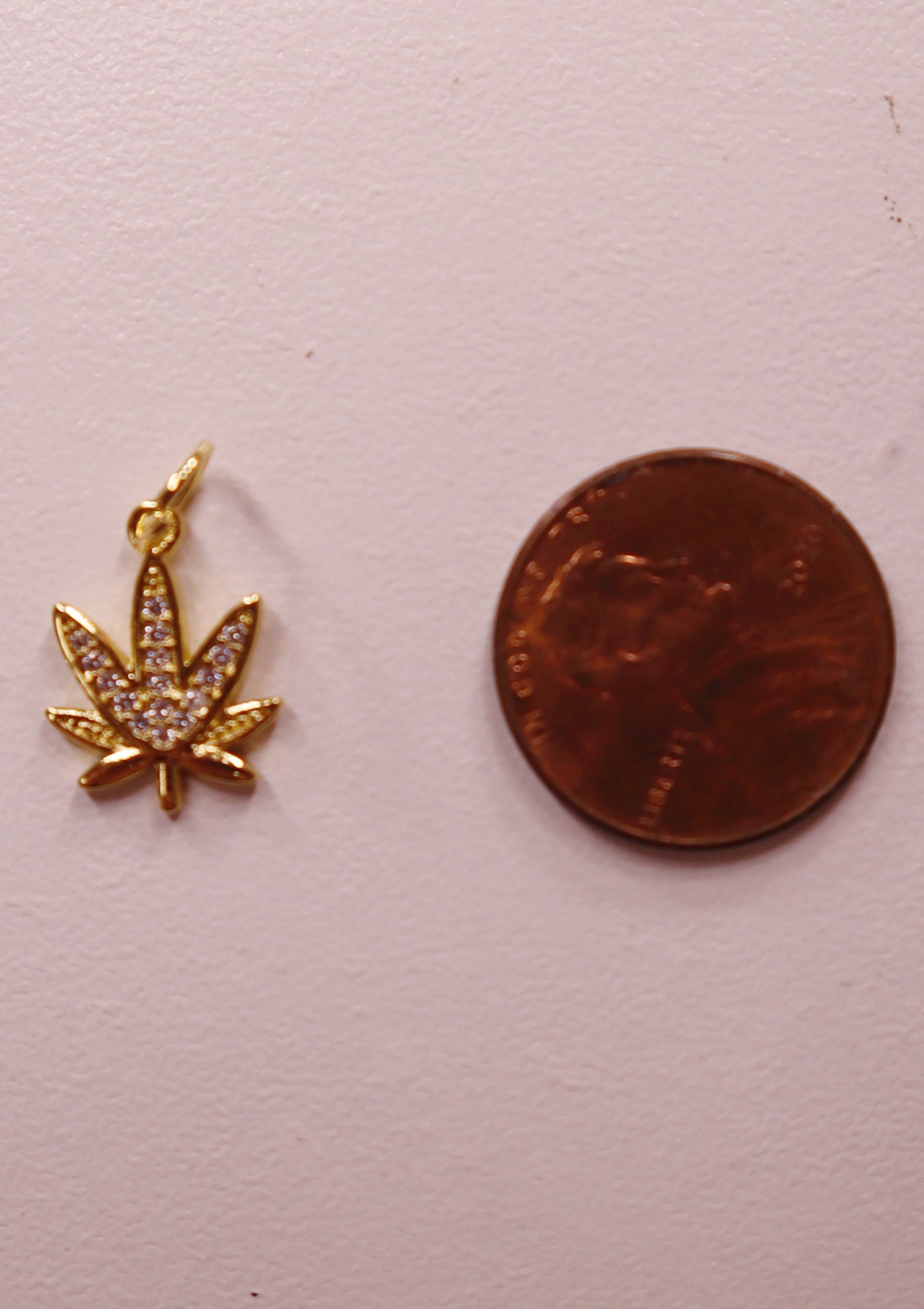 Gold marijuana leaf charm next to a penny on a light pink background