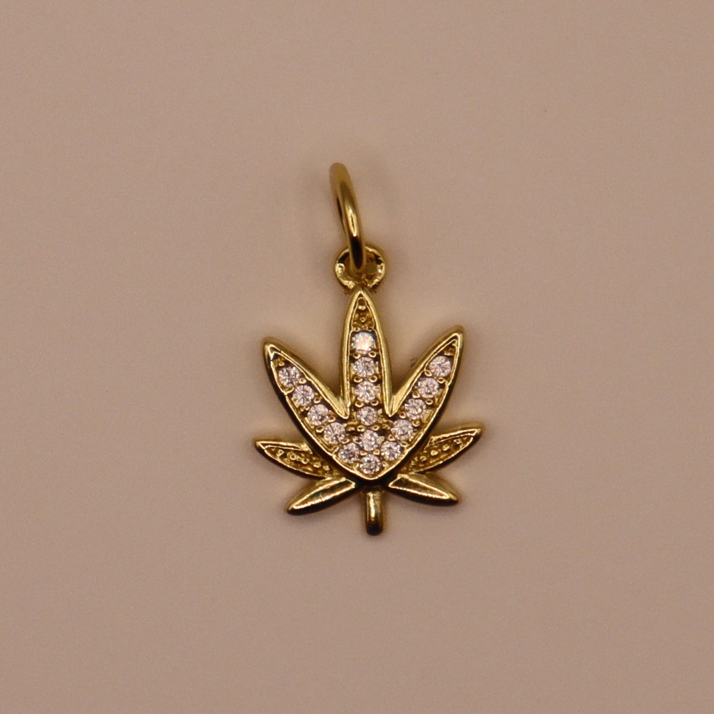 Gold pendant shaped like a cannabis leaf with embedded stones on a beige background