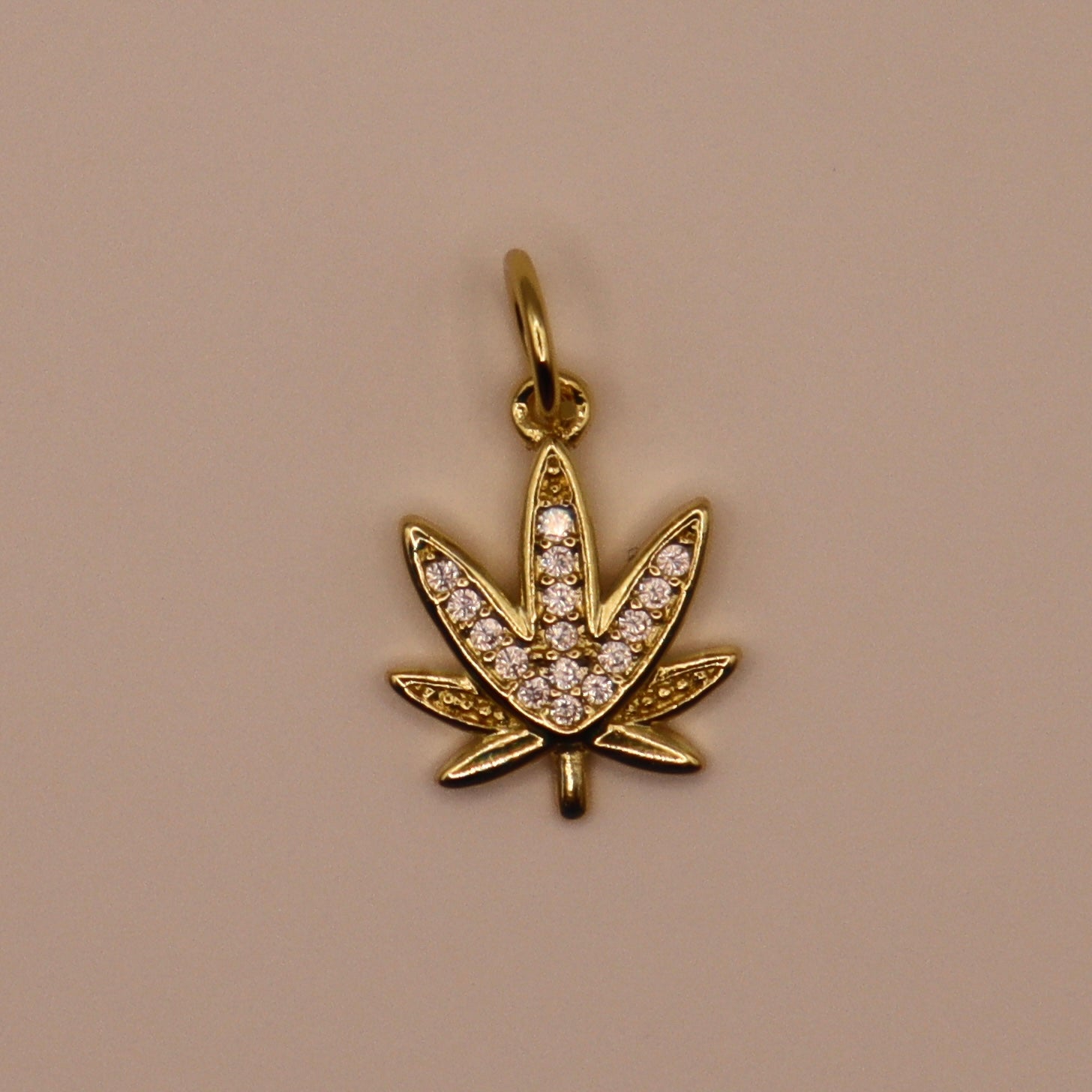 Gold pendant shaped like a cannabis leaf with embedded stones on a beige background