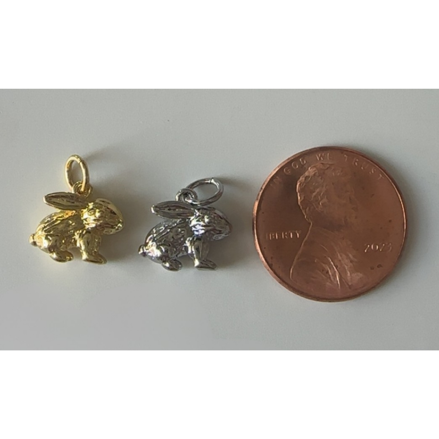 Two rabbit-shaped charms, one gold and one silver, next to a penny for size comparison on a white background.
