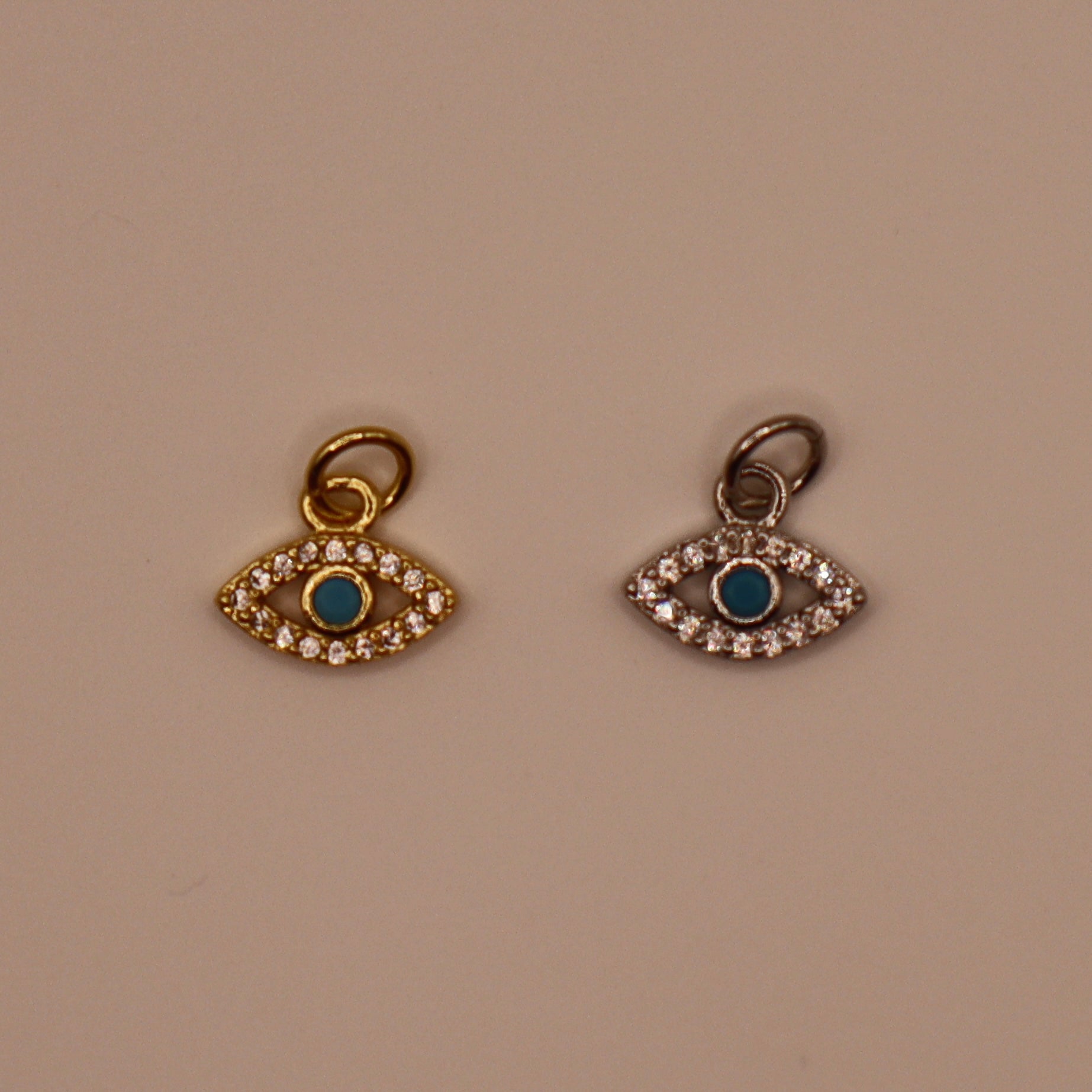 Two evil eye charms with blue centers and gold and silver borders on a beige background.