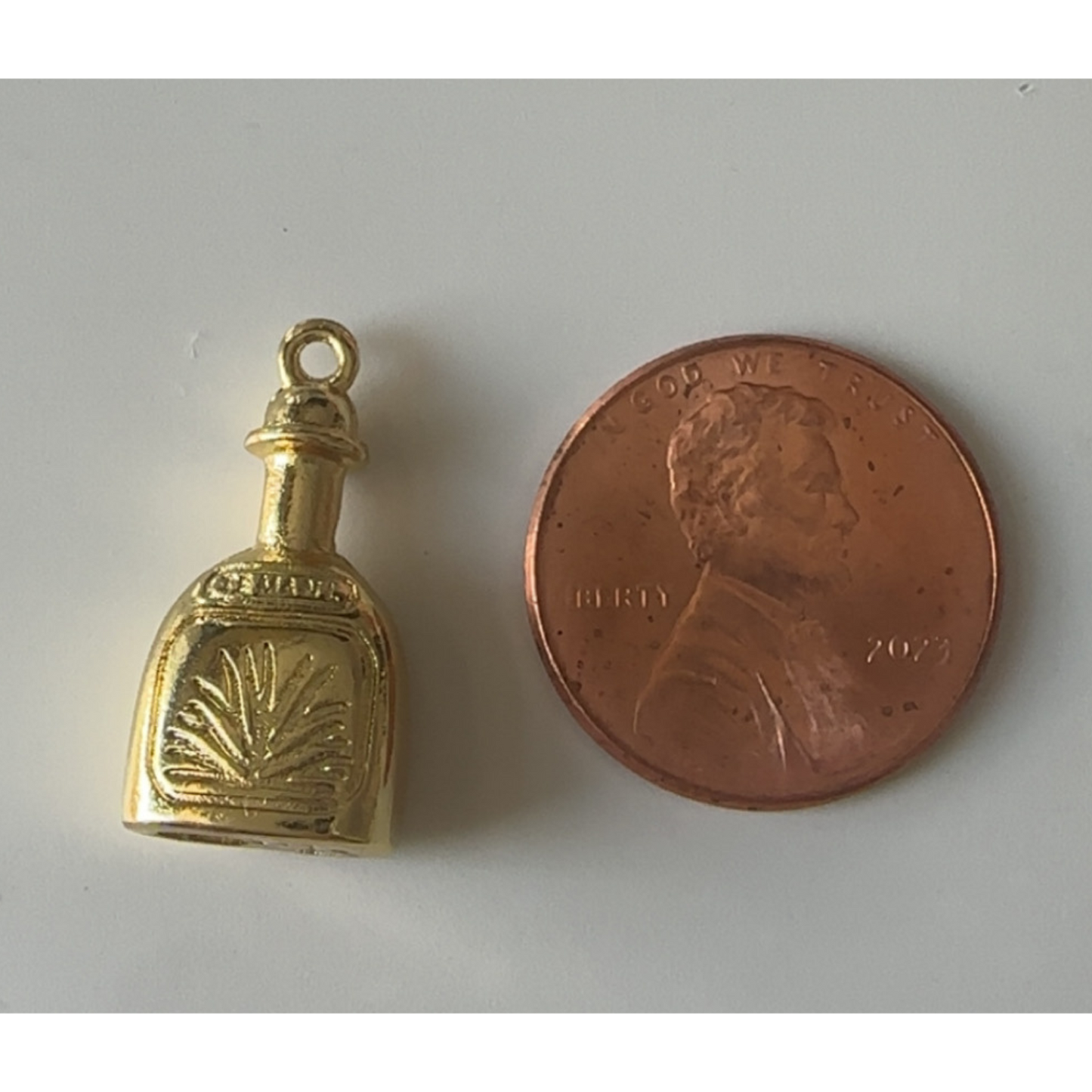 Gold tequila charm next to a penny on a white background