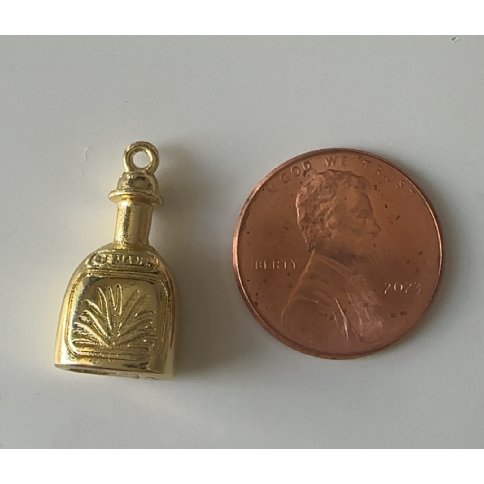 Gold tequila charm next to a penny on a white background