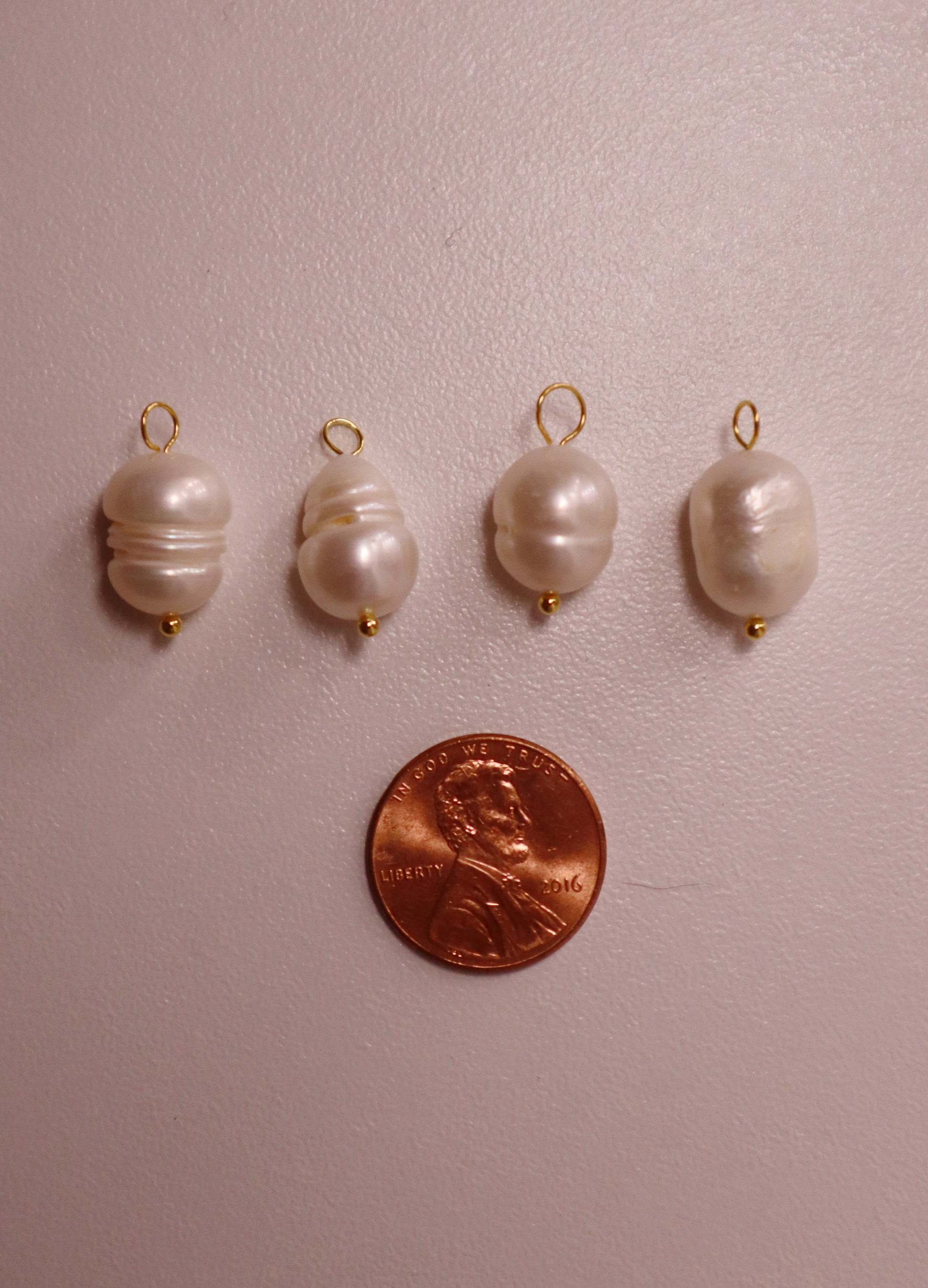 Four pearl pendants with gold clasps on a pink surface, next to a penny for scale.
