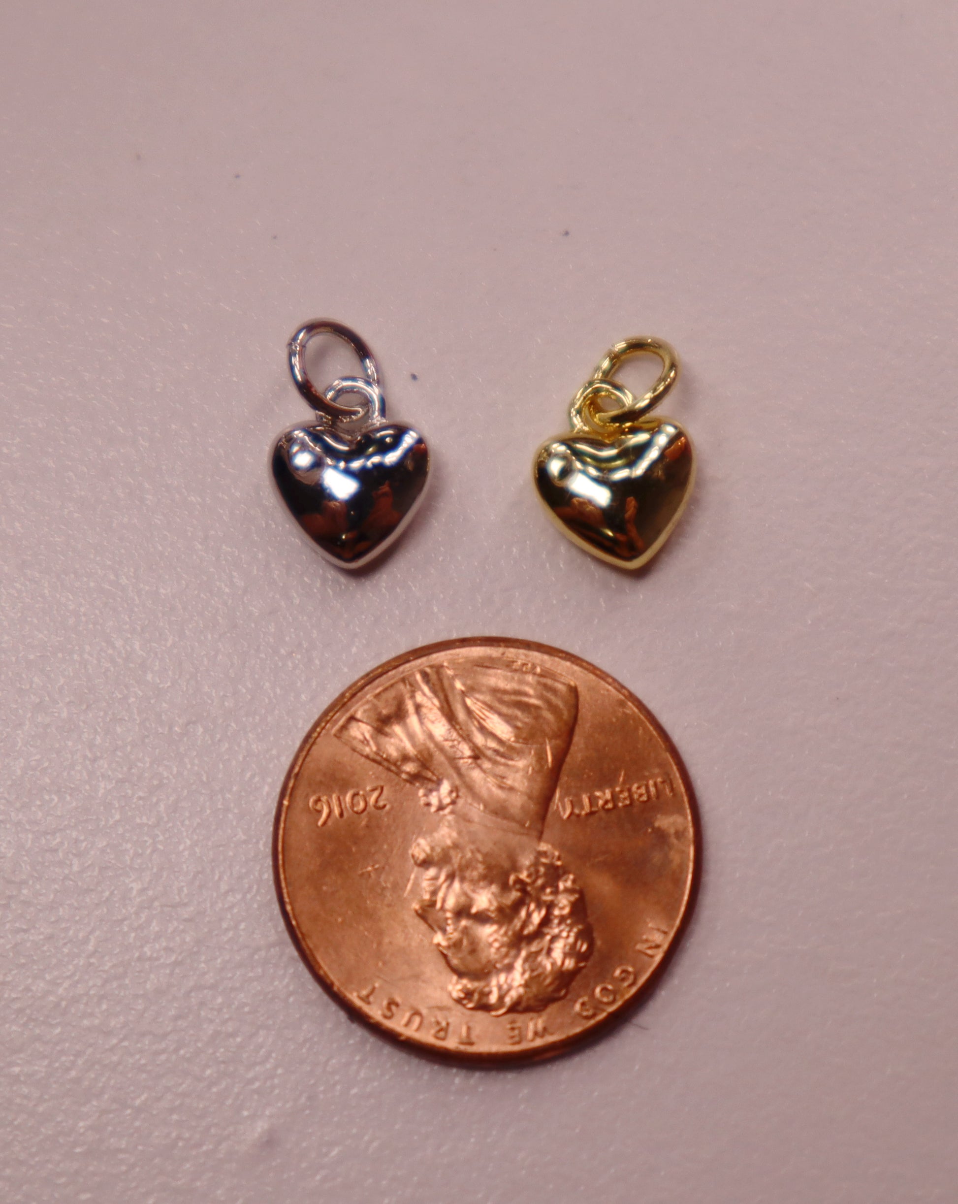 Two heart-shaped charms, one silver and one gold, next to a penny on a pink surface.