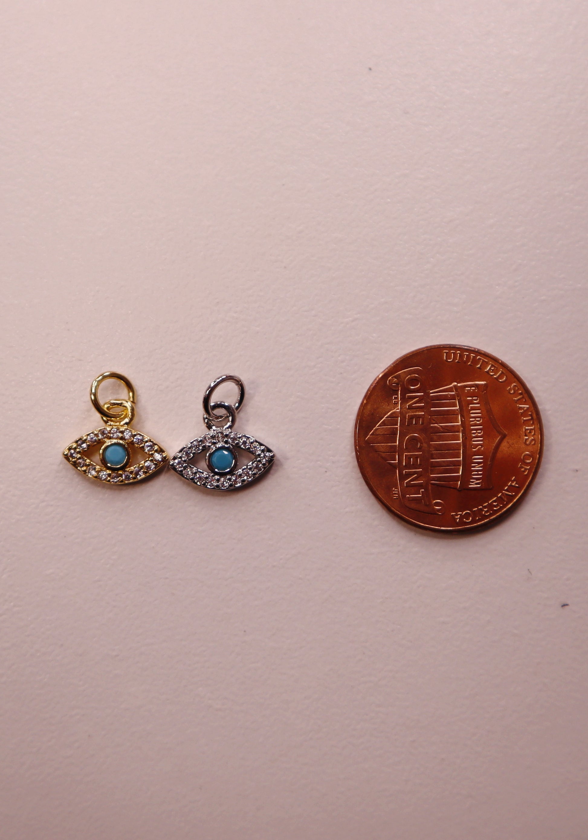 Two evil eye-shaped charms with turquoise stones next to a penny on a light pink background