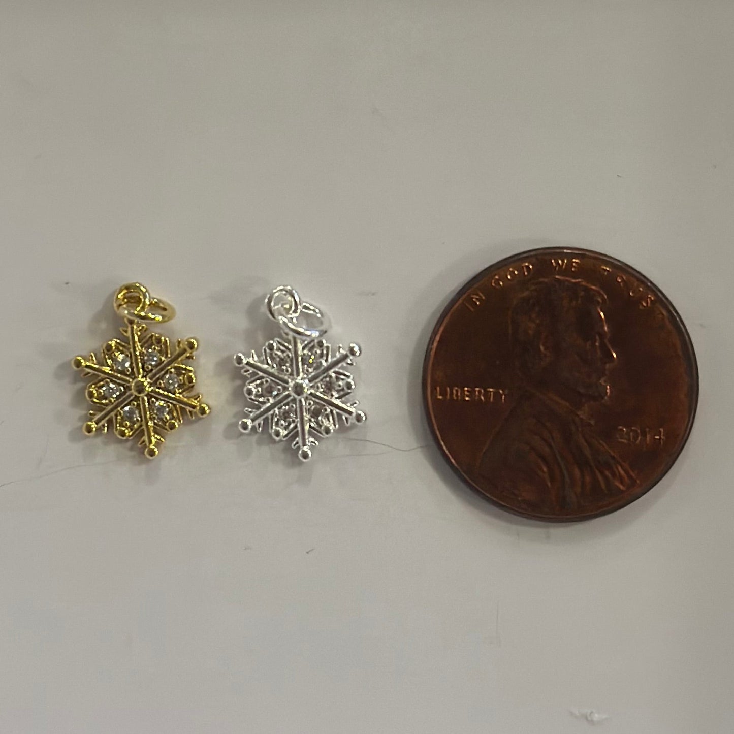 Two snowflake-shaped charms, one gold and one silver, next to a penny on a white background.